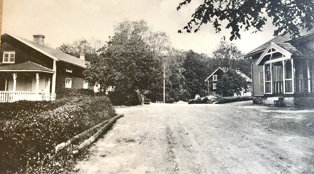 Svartvit foto med en väg i mitten som omges av röda hus. Till vänster syns ett hus med en veranda.